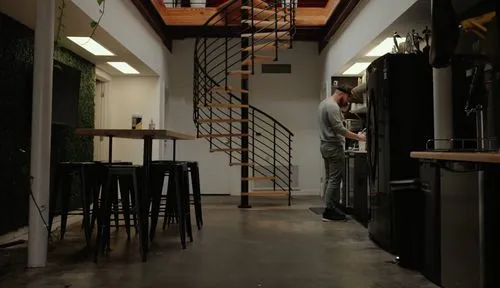 Jeremy stands by a kitchen area in a modern office with a spiral staircase and high tables with stools.