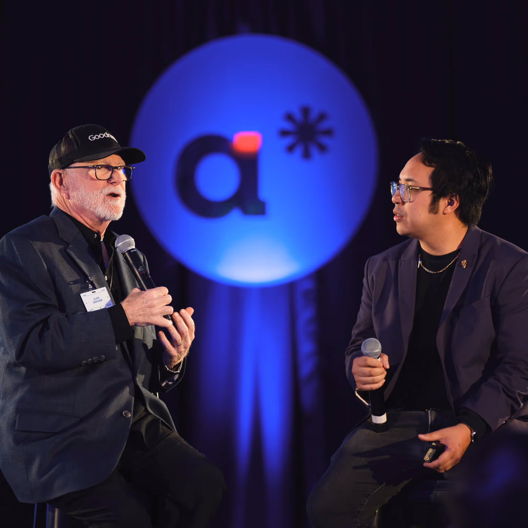 Two men seated and holding microphones, engaged in a conversation on stage with a blue lit circular logo behind them.