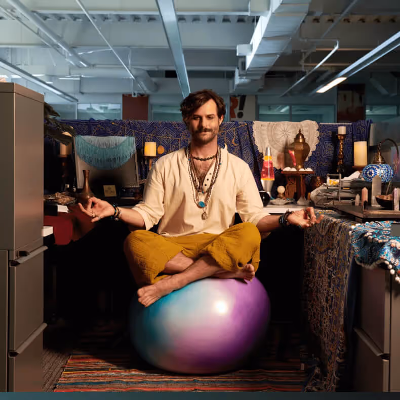 Man wearing yellow pants and a beige shirt meditation-cross-legged on a colorful exercise ball in an office cubicle decorated with candles and fabrics.