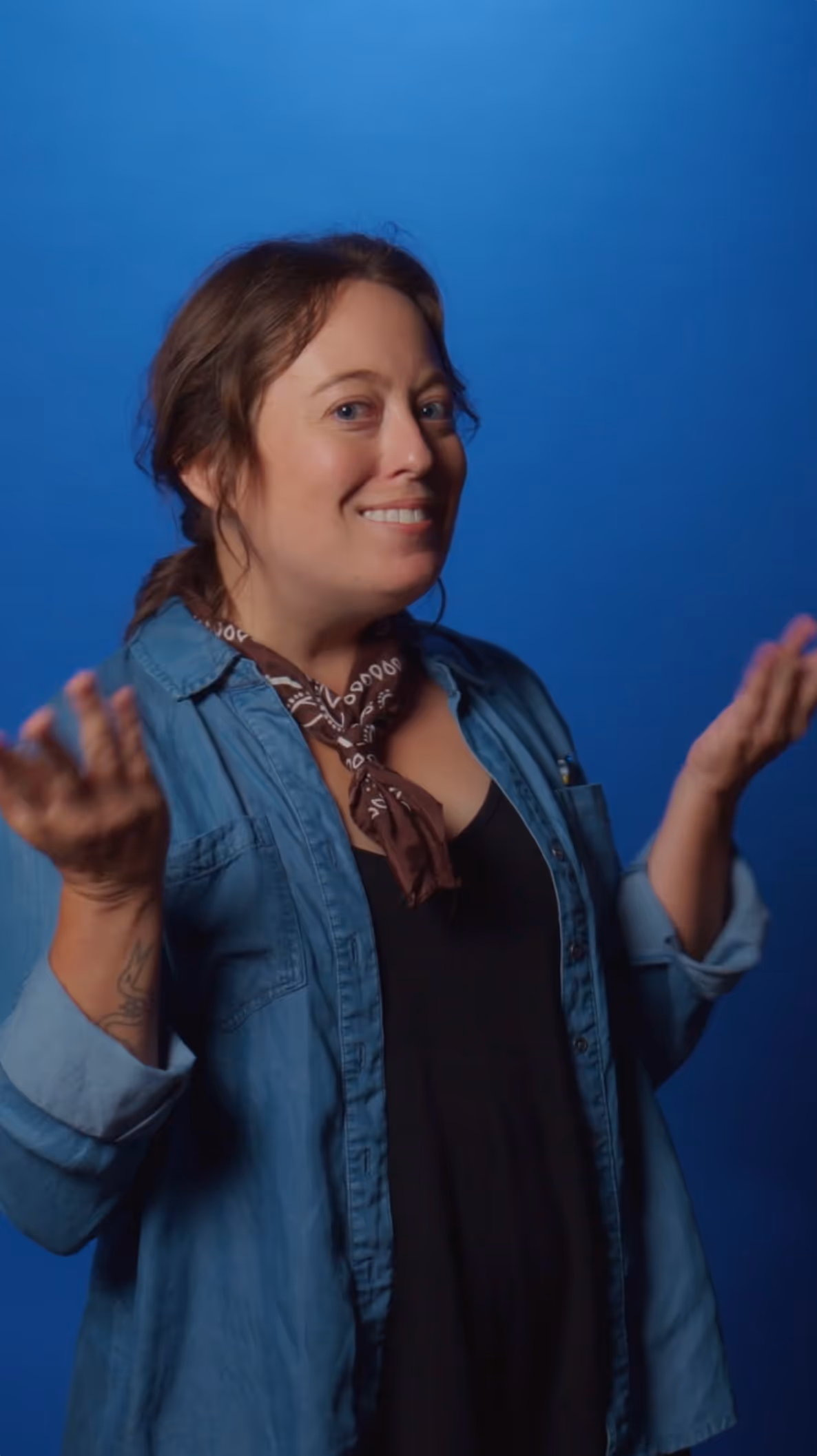 Smiling woman in a denim shirt and brown scarf gesturing with both hands against a blue background.