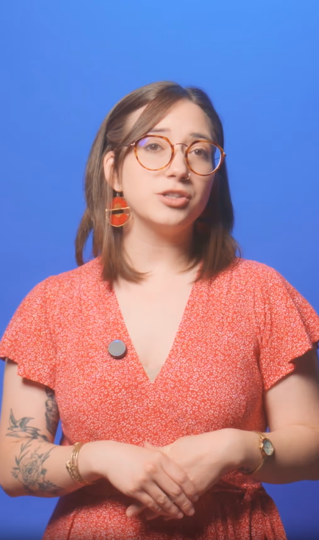 Woman with shoulder-length brown hair, round glasses, and red floral dress speaking against a blue background.