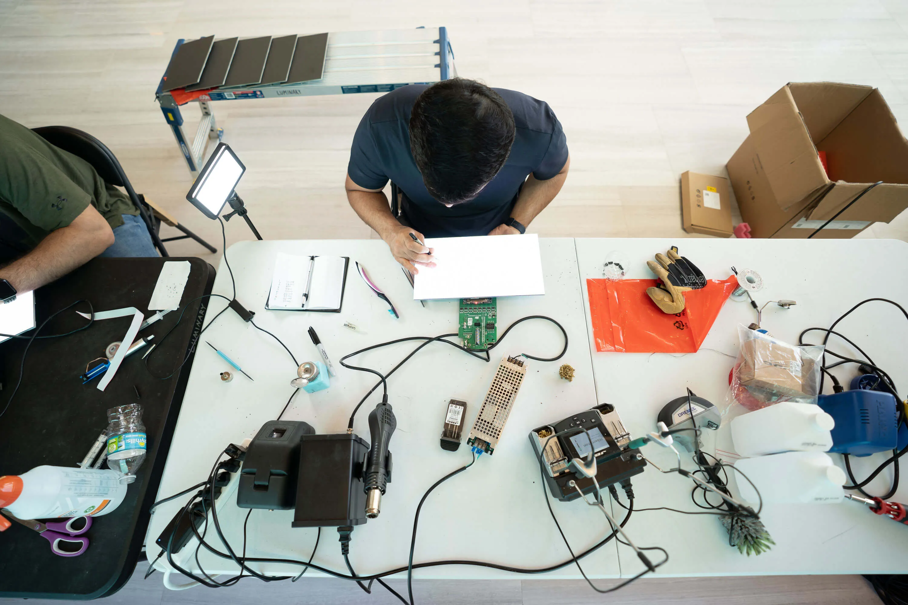 Overhead view of a person working at a cluttered white table with electronic tools, circuit boards, gloves, and other equipment.