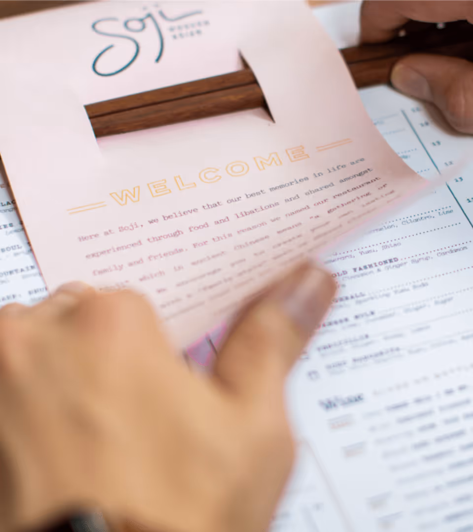 Close-up of hands holding a pink Soji brand menu with a 'WELCOME' message above the menu items in the background.