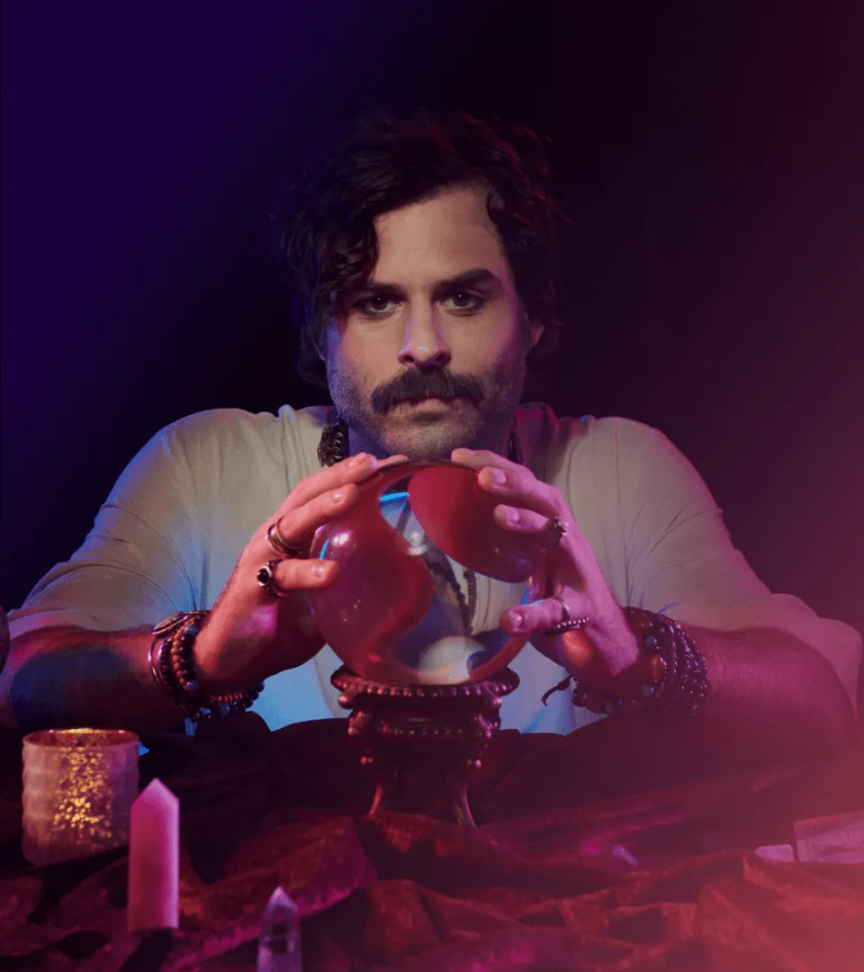 Man with a mustache and curly hair gazing into a crystal ball with focused expression.