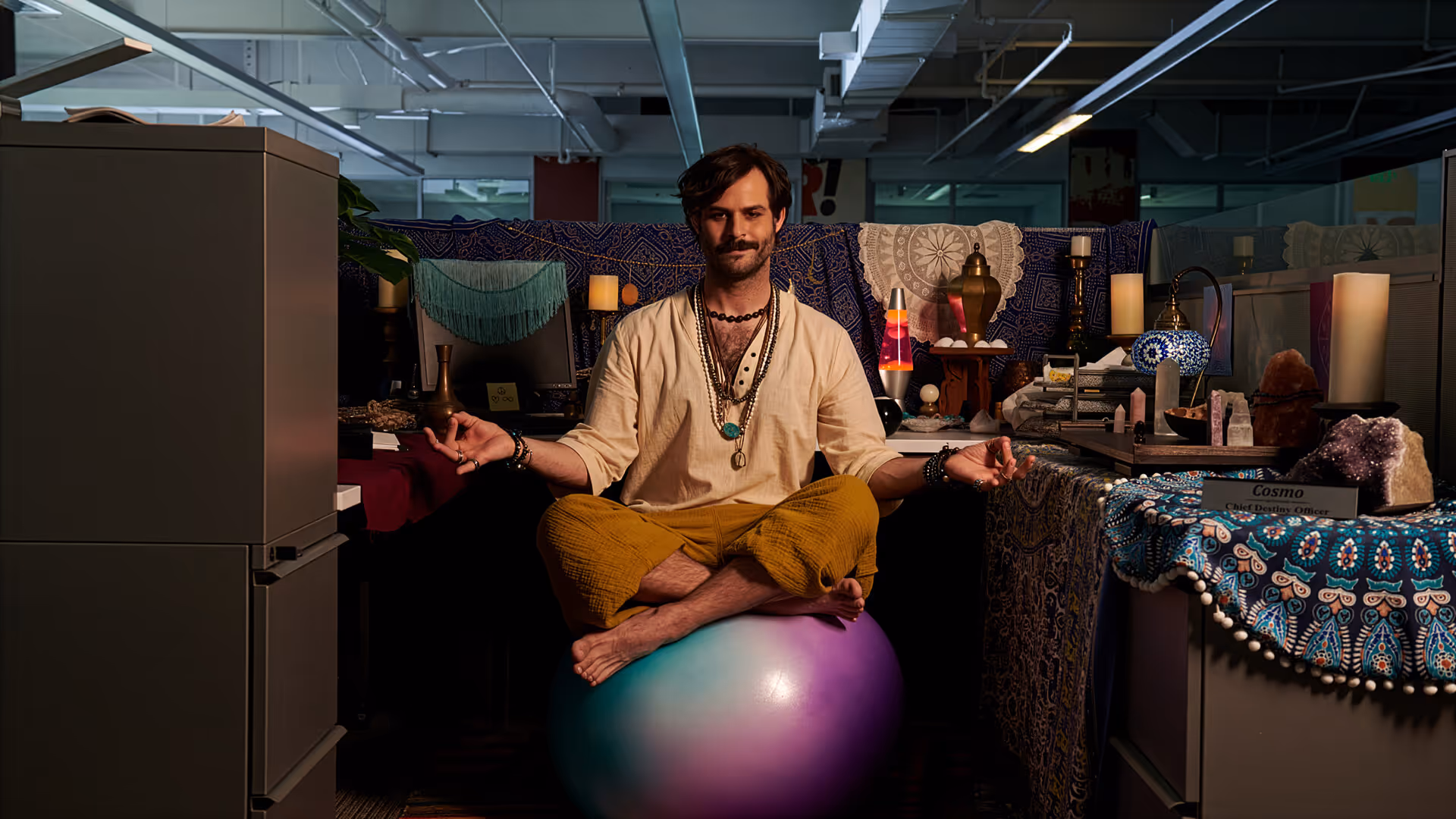 Man in casual clothes sitting cross-legged on a colorful exercise ball meditating in a decorated office cubicle.