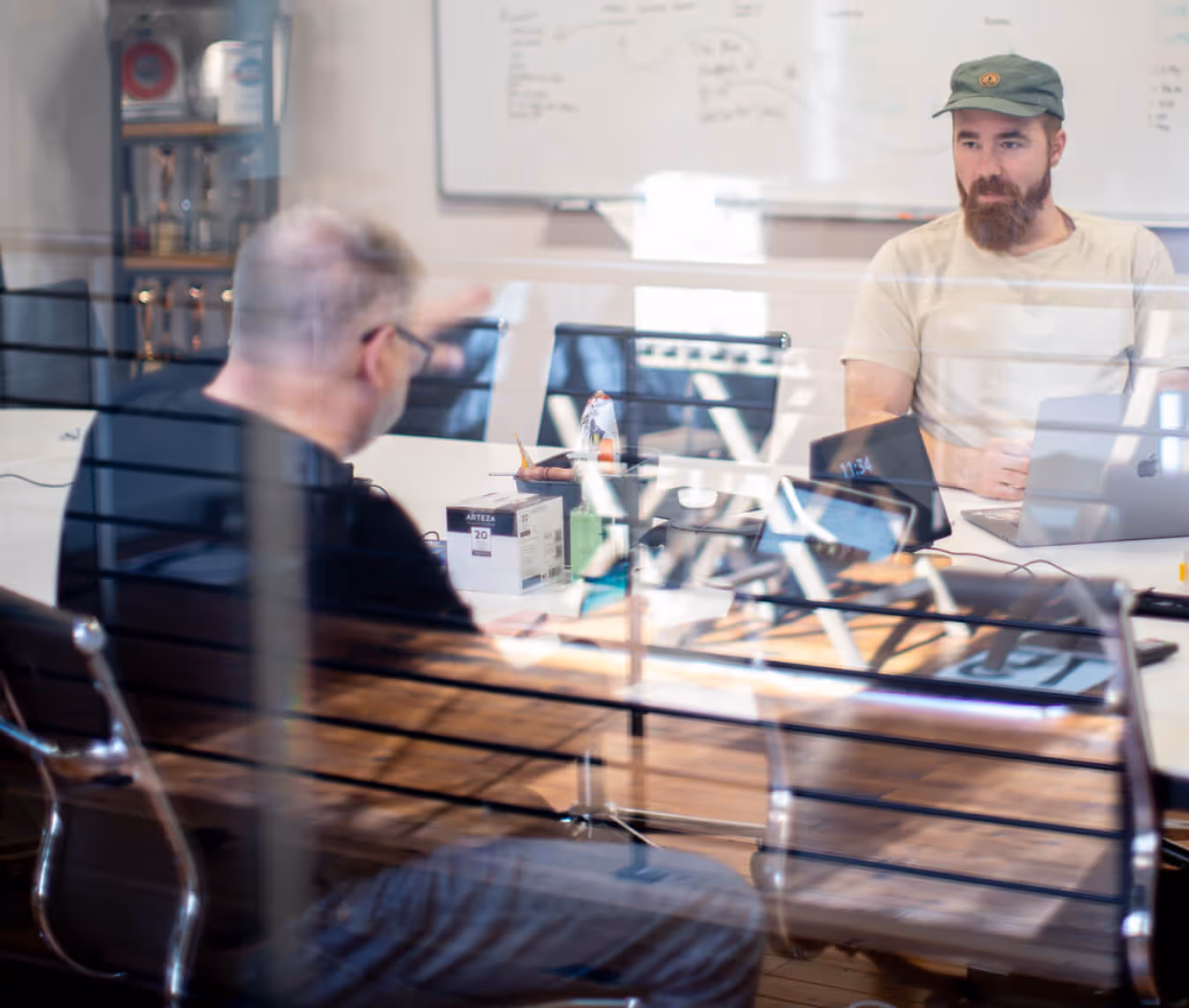 Two men having a discussion across a conference table with laptops and office supplies, seen through a glass partition.