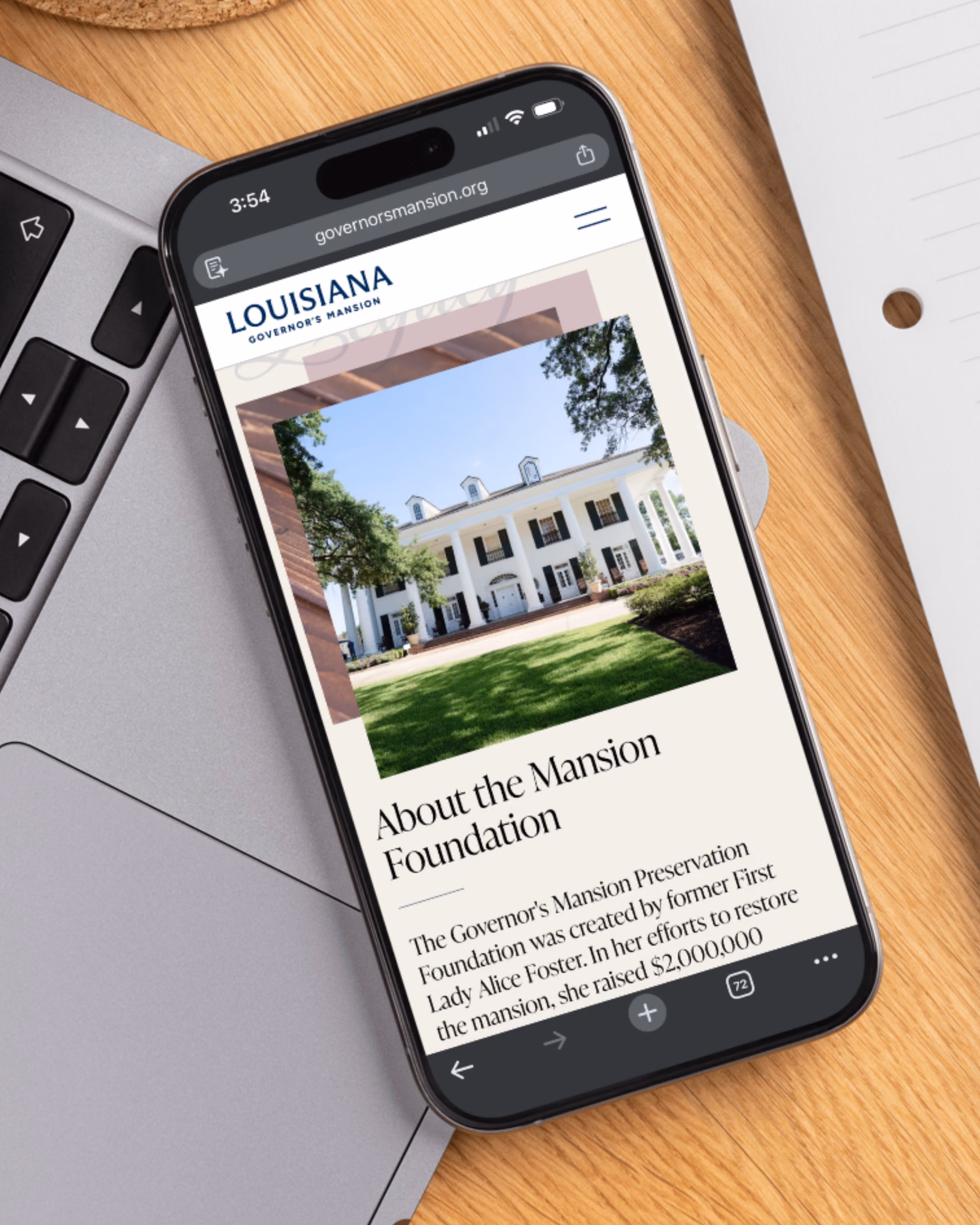 Smartphone on a wooden desk displaying the Louisiana Governor's Mansion website with an image of the mansion and text about the Mansion Foundation.