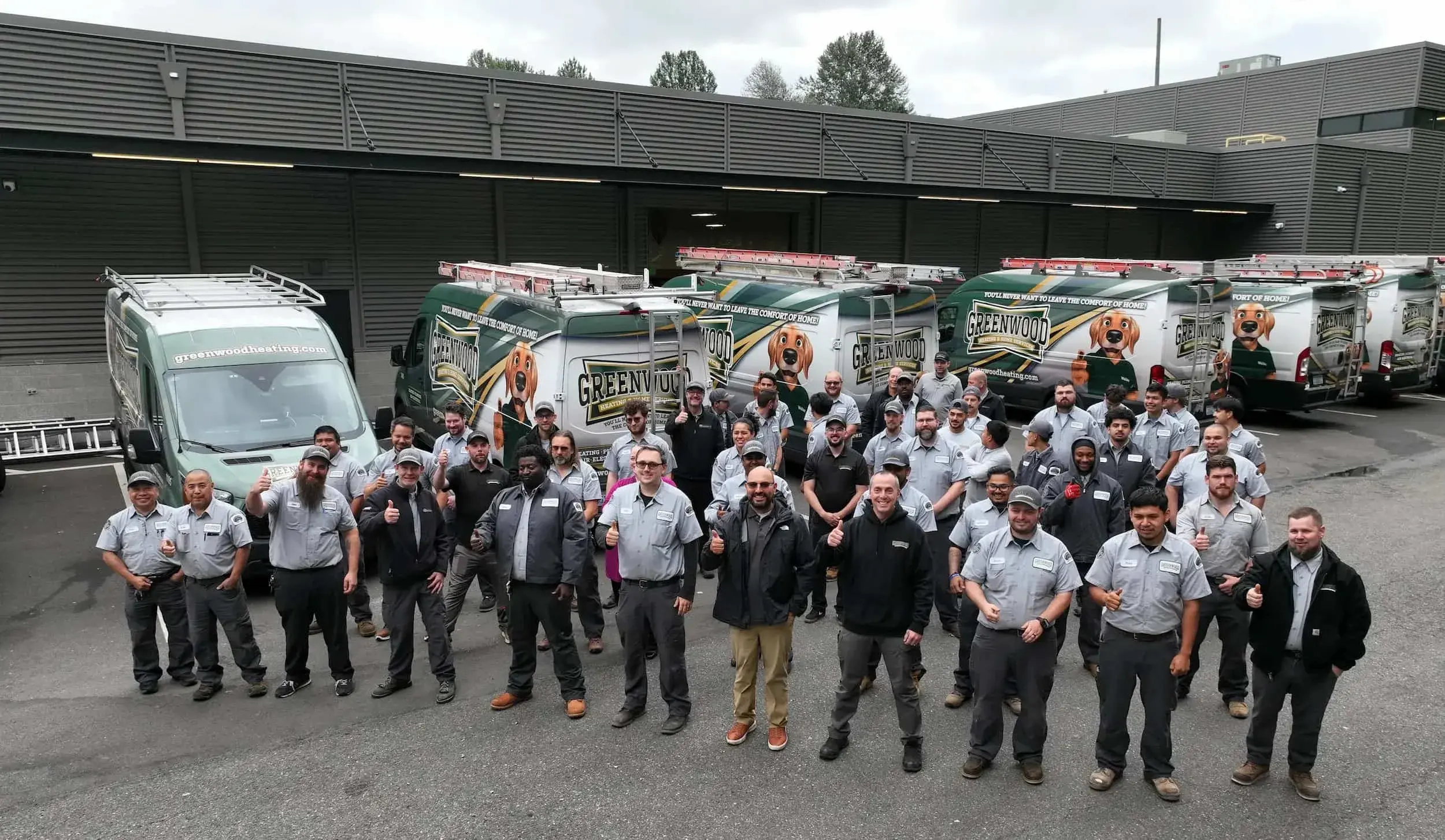 Group of men and women posing outdoors in uniforms in front of several Greenwood Heating company service vans with logo and cartoon dog on them.