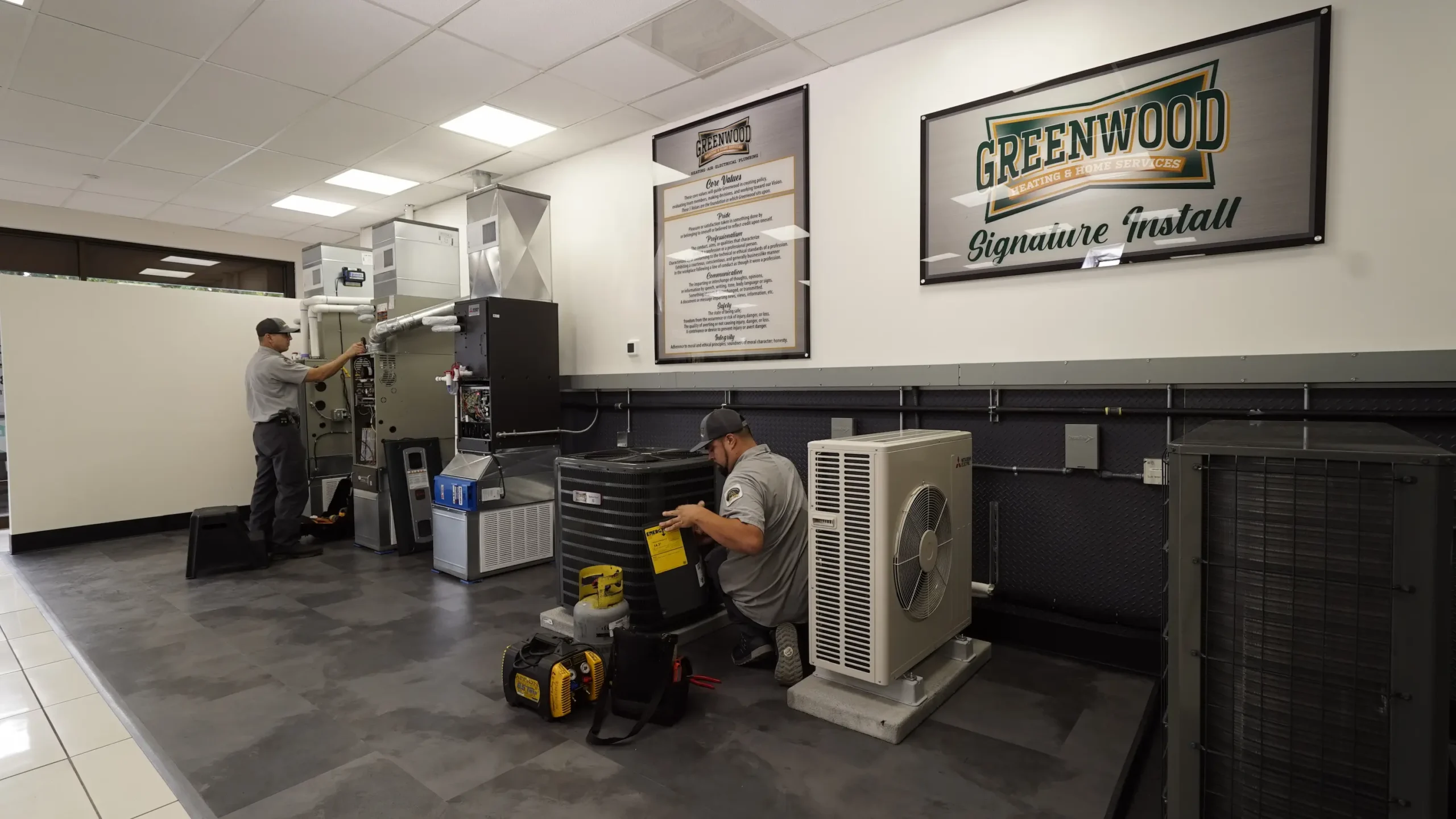Two technicians in gray uniforms working on HVAC units inside a clean training facility with 'Greenwood Signature Install' and company core values framed on the wall.