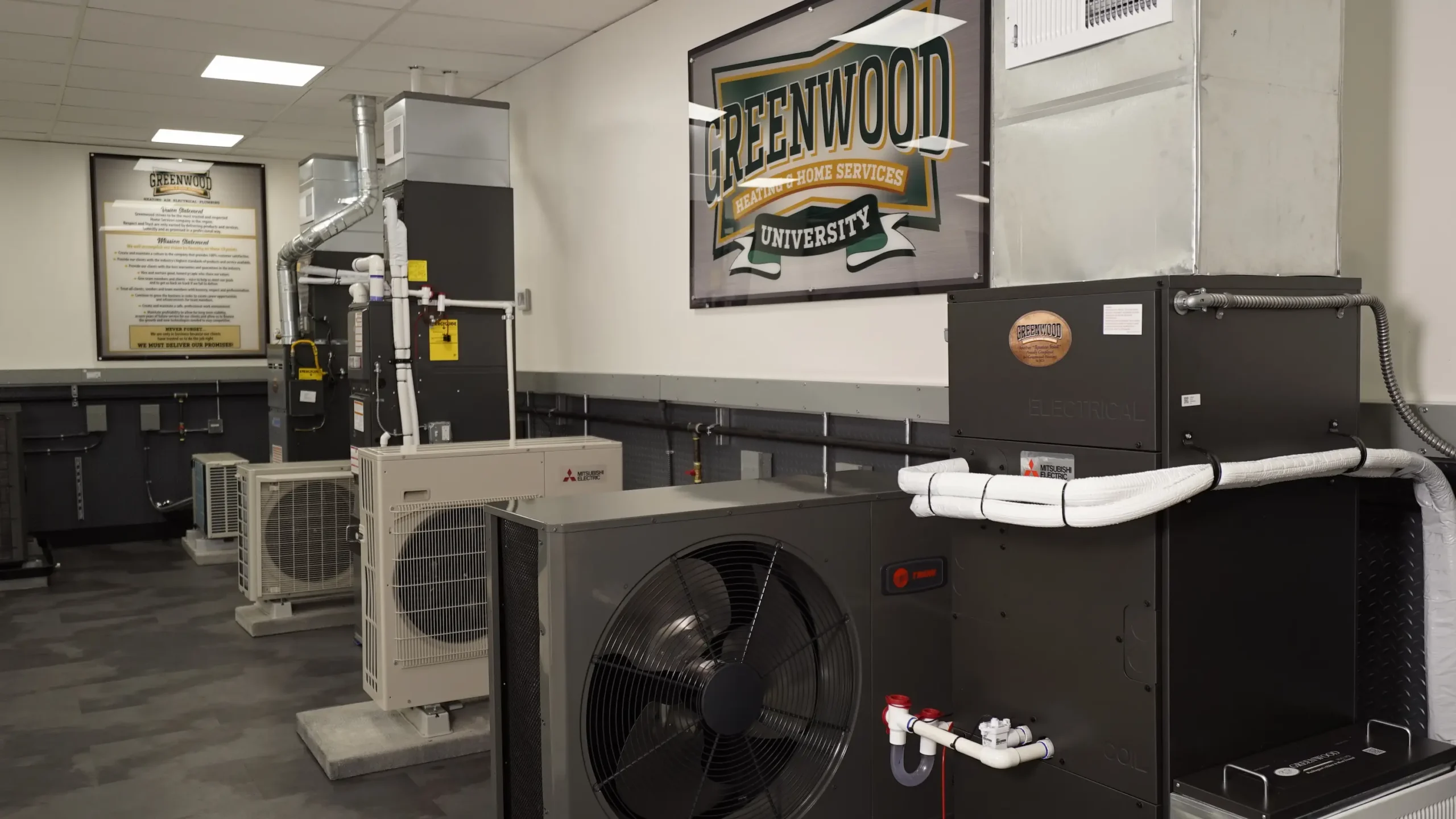 Indoor training room with multiple HVAC units and equipment lined up against a wall, with a Greenwood University Heating & Home Services sign displayed prominently on the wall above the units.