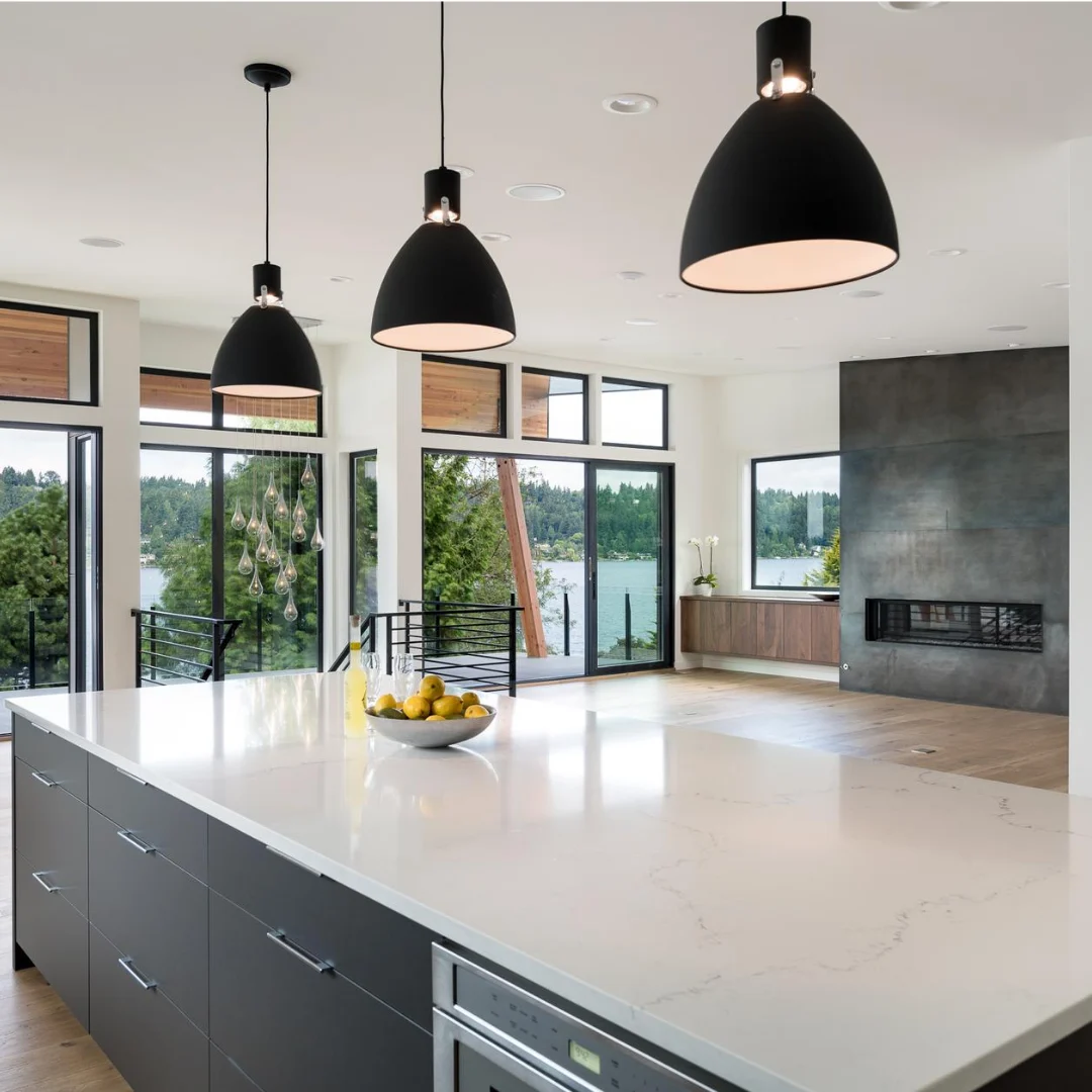 Modern kitchen interior with a large white marble island countertop and dark cabinets, three black pendant lights hanging above, and glass doors and windows showing a lake and trees outside, with a built-in fireplace and wooden flooring in the background.
