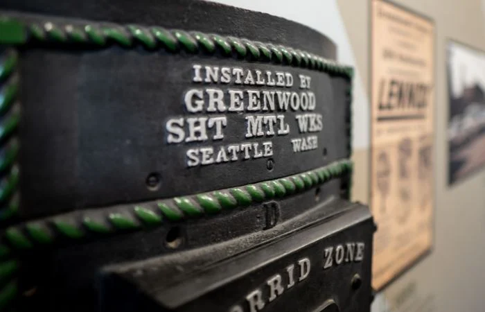 Close-up of a metal plaque on a device reading INSTALLED BY GREENWOOD SHT MTL WKS SEATTLE WASH with green rope-like trim, with blurred posters in the background.