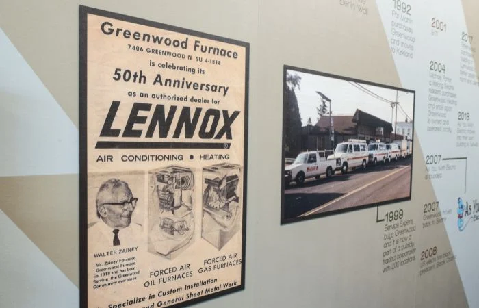 A timeline display on a wall featuring a vintage Greenwood Furnace advertisement celebrating its 50th anniversary as an authorized dealer for Lennox air conditioning and heating, alongside a photo of a fleet of white service vans parked on a street.