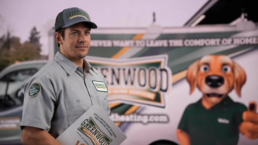 Man in a Greenwood company uniform holding a Greenwood brochure, standing in front of a company van with Greenwood branding and a cartoon dog mascot.