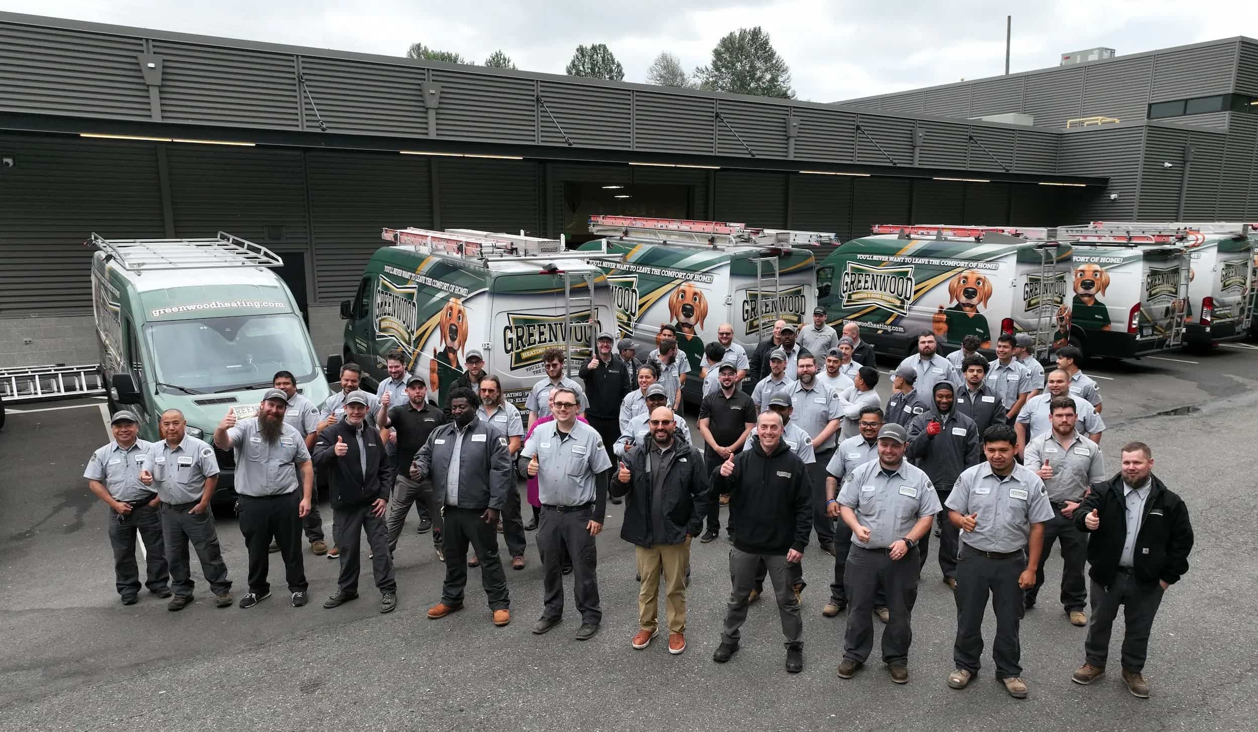 Group of workers standing in front of several Greenwood Heating service vans, giving thumbs up in an outdoor parking area next to a building.