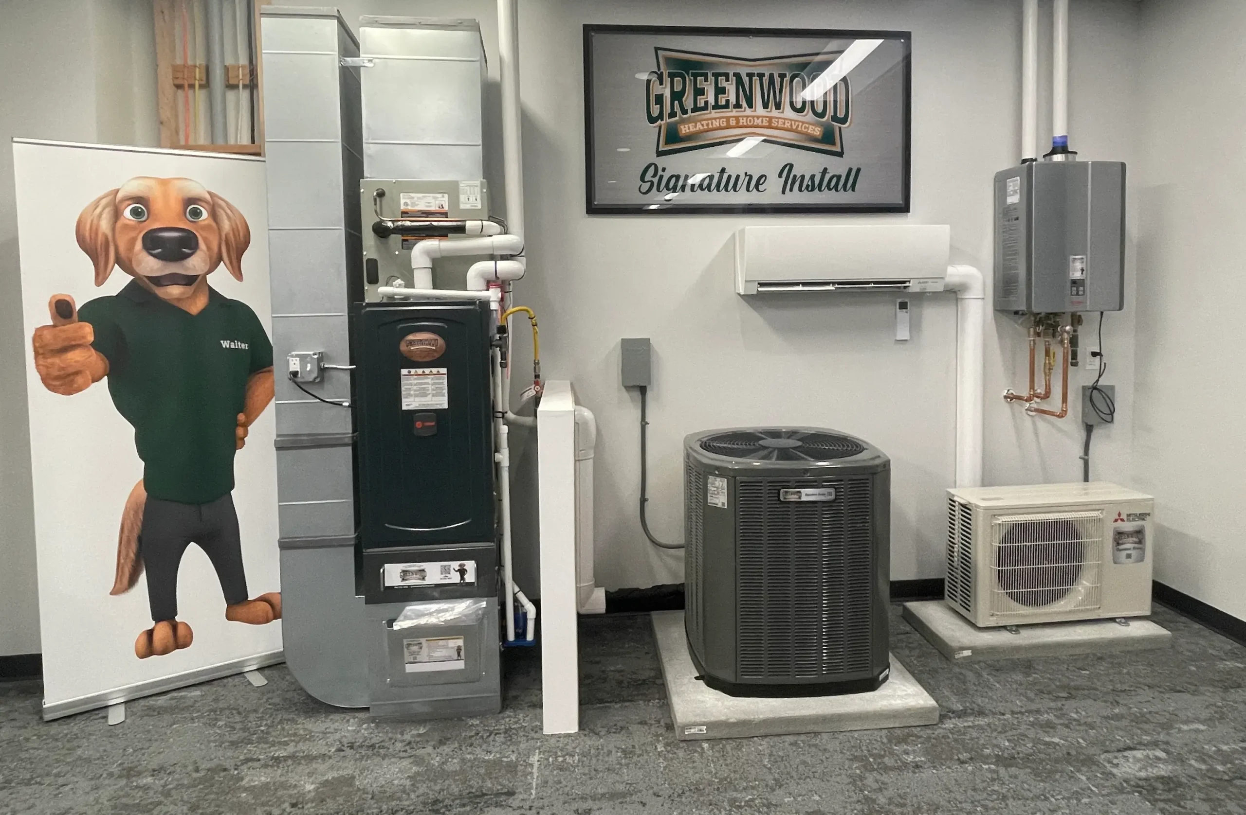 Indoor showroom display featuring HVAC equipment including a heating furnace, air conditioning units, and a water heater, with a banner of an anthropomorphic dog character named Walter giving a thumbs up on the left and a framed sign reading 'Greenwood Heating & Home Services Signature Install' on the wall.