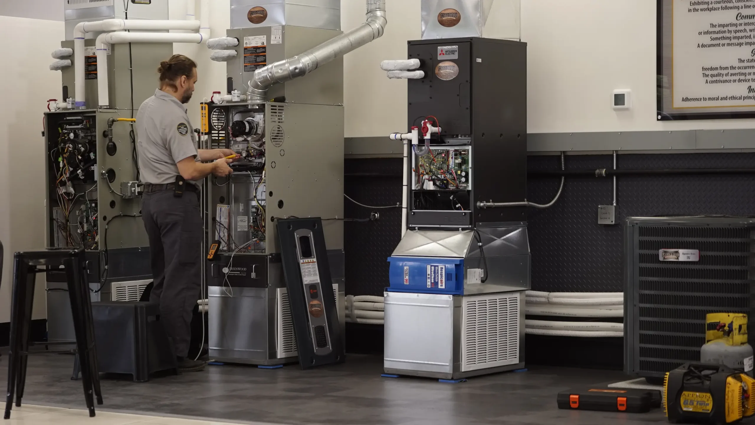 A technician with a beard and tied-back hair works on the inside components of a large HVAC unit in a workshop, surrounded by various equipment and machinery panels.