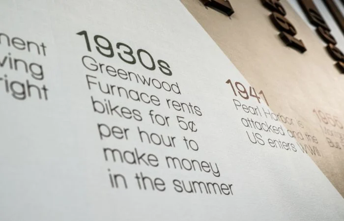 Text on a wall showing a timeline entry for the 1930s stating 'Greenwood Furnace rents bikes for 5¢ per hour to make money in the summer.'