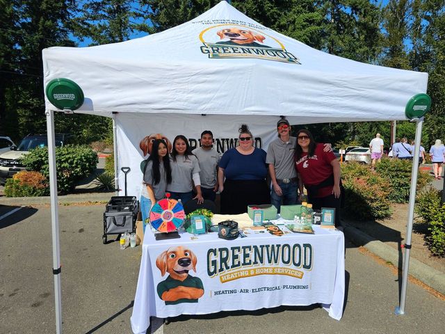 Six people standing behind a promotional booth under a white canopy tent with Greenwood Heating & Home Services branding, including a logo of a cartoon dog, with various items displayed on the table.