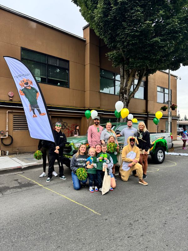 Group of adults and children posing in front of a decorated pickup truck with green, white, and yellow balloons; one person is holding a vertical flag with a cartoon dog, and another is dressed in a dog costume.