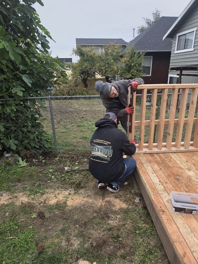 Two men working together to install a wooden railing on a deck in a backyard, one holding the railing upright while the other secures it at the base.