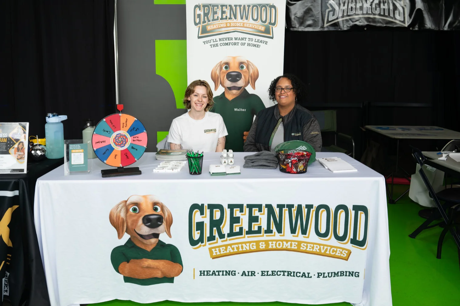 Two women sitting behind a Greenwood Heating & Home Services booth with promotional items including a prize wheel, pens, hand sanitizer, and brochures, against a backdrop featuring a cartoon dog mascot named Walter.