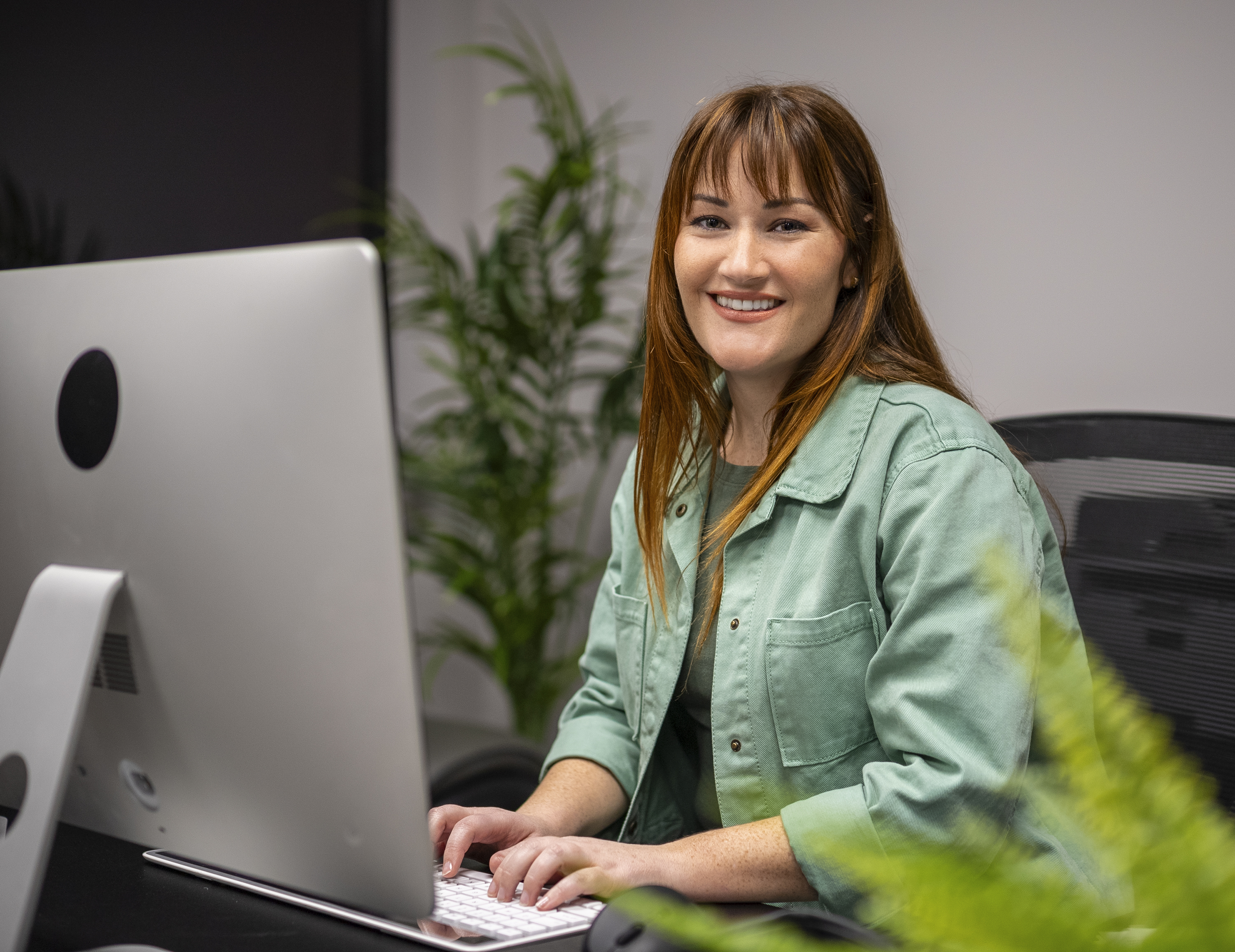 Smiling woman with red hair, wearing a green jacket, typing on a keyboard in front of a large desktop computer in an office with plants.
