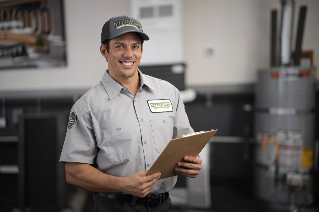 Smiling man in a Greenwood uniform and cap holding a clipboard, standing indoors near a water heater.