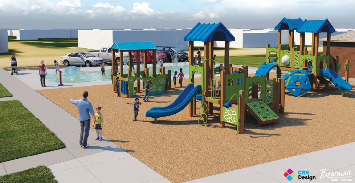 Playground with blue and green climbing structures and slides surrounded by children playing and adults supervising on a sandy area, with a splash pad area and parked cars in the background.