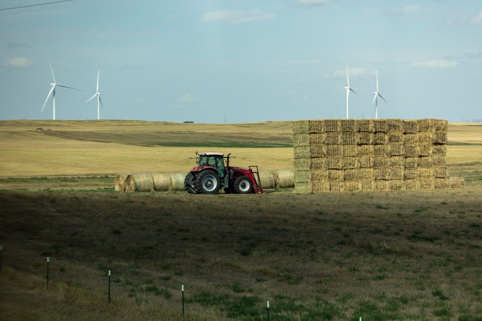 Red tractor parked in a field next to a large stack of square hay bales with round hay bales lined up in a row behind it, and four wind turbines in the distance under a partly cloudy sky.