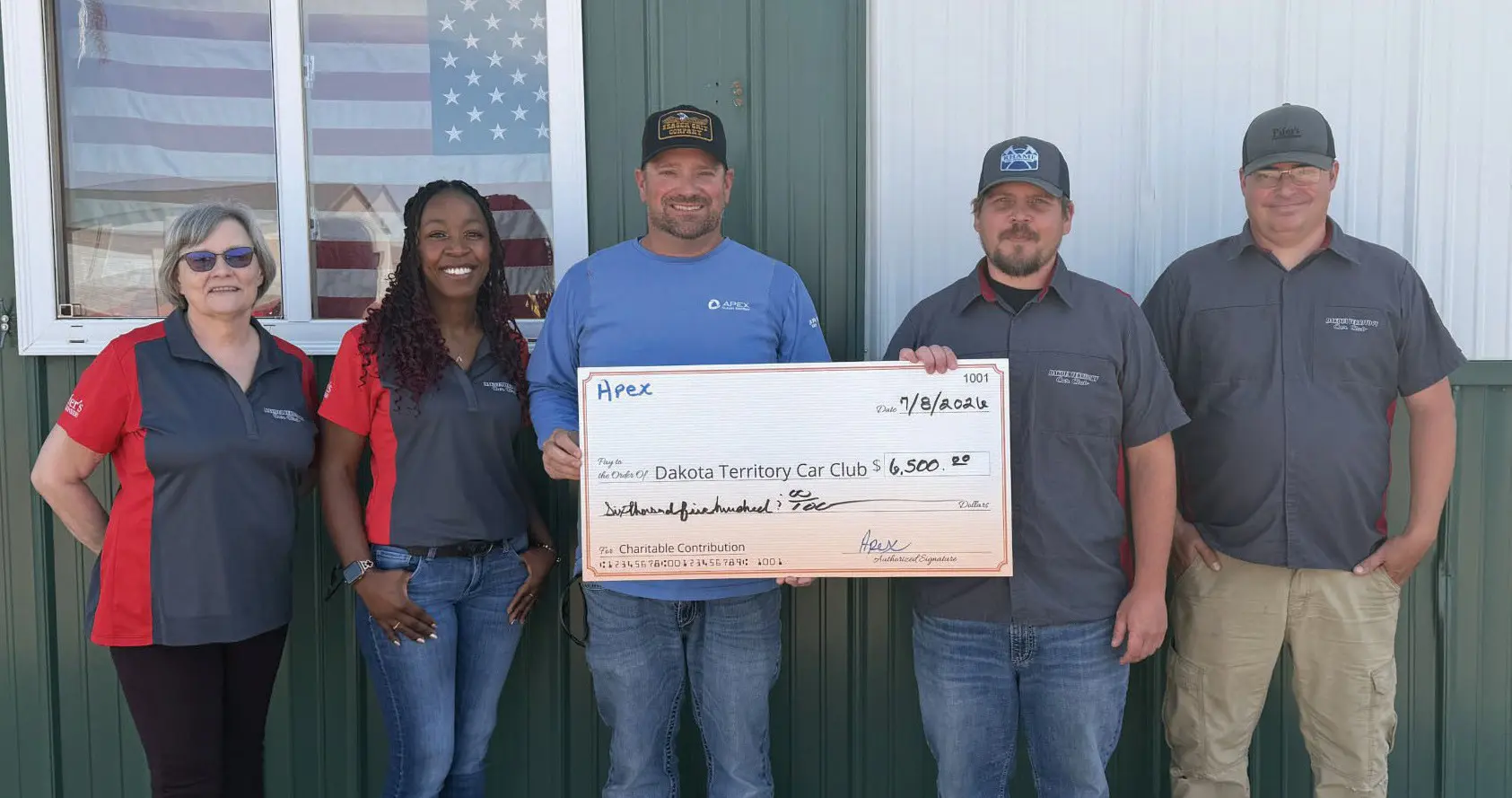 Five people standing in a row in front of a green wall with an American flag in a window, two men in the center holding a large check for $6,500 payable to Dakota Territory Car Club dated 7/8/2021.
