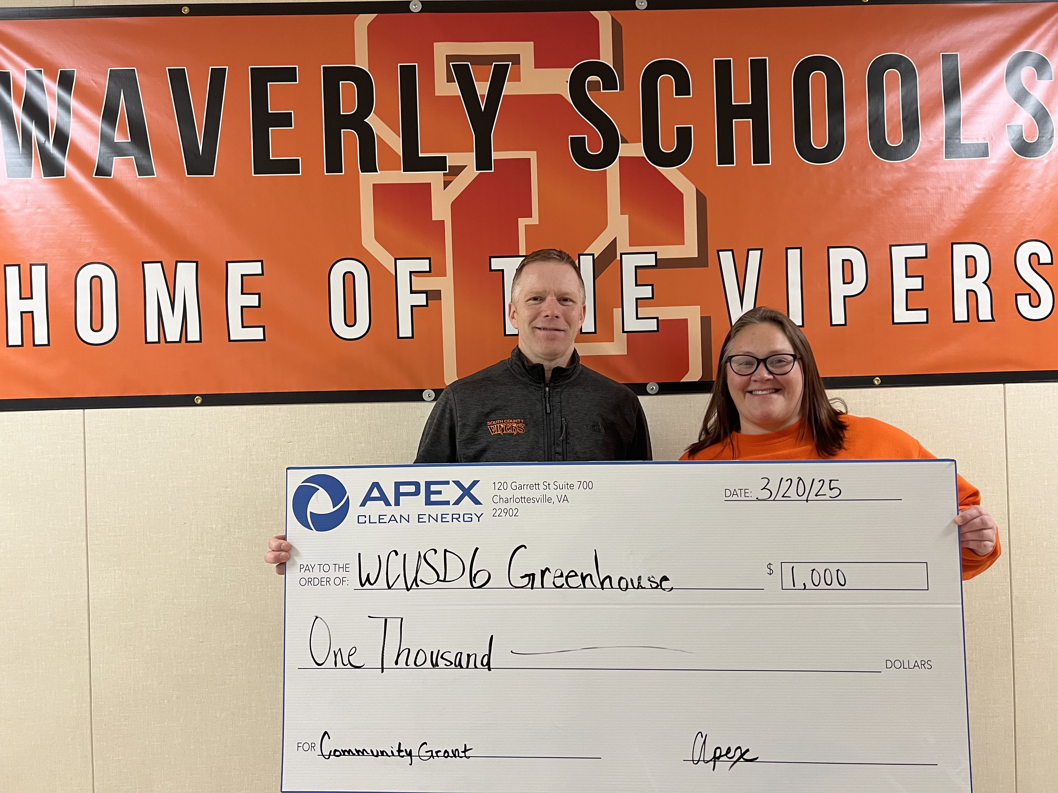 Two people standing indoors in front of an orange Waverly Schools banner that reads 'Home of the Vipers' holding a large check from Apex Clean Energy for one thousand dollars, dated 3/20/25, payable to WCUSD6 Greenhouse for a community grant.