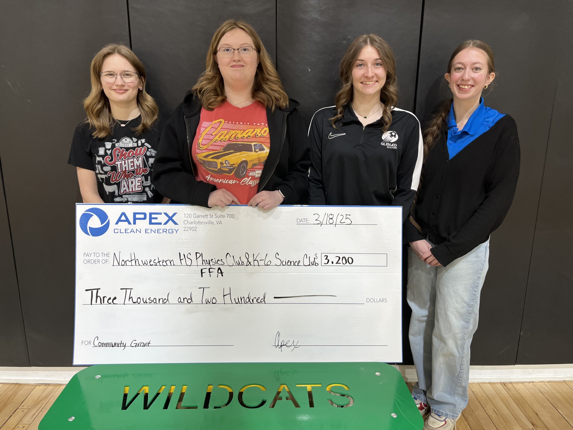 Four young women posing indoors holding a large check from Apex Clean Energy for $3,200 made out to Northwestern HS Physics Club, K-6 Science Club, and FFA, dated 3/18/25 for a community grant, standing behind a green bench with the word WILDCATS cut out.
