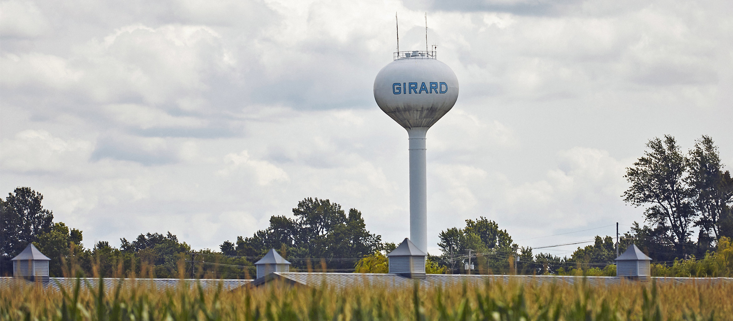 Large water tower labeled Girard standing behind trees and buildings with cornfield in foreground.