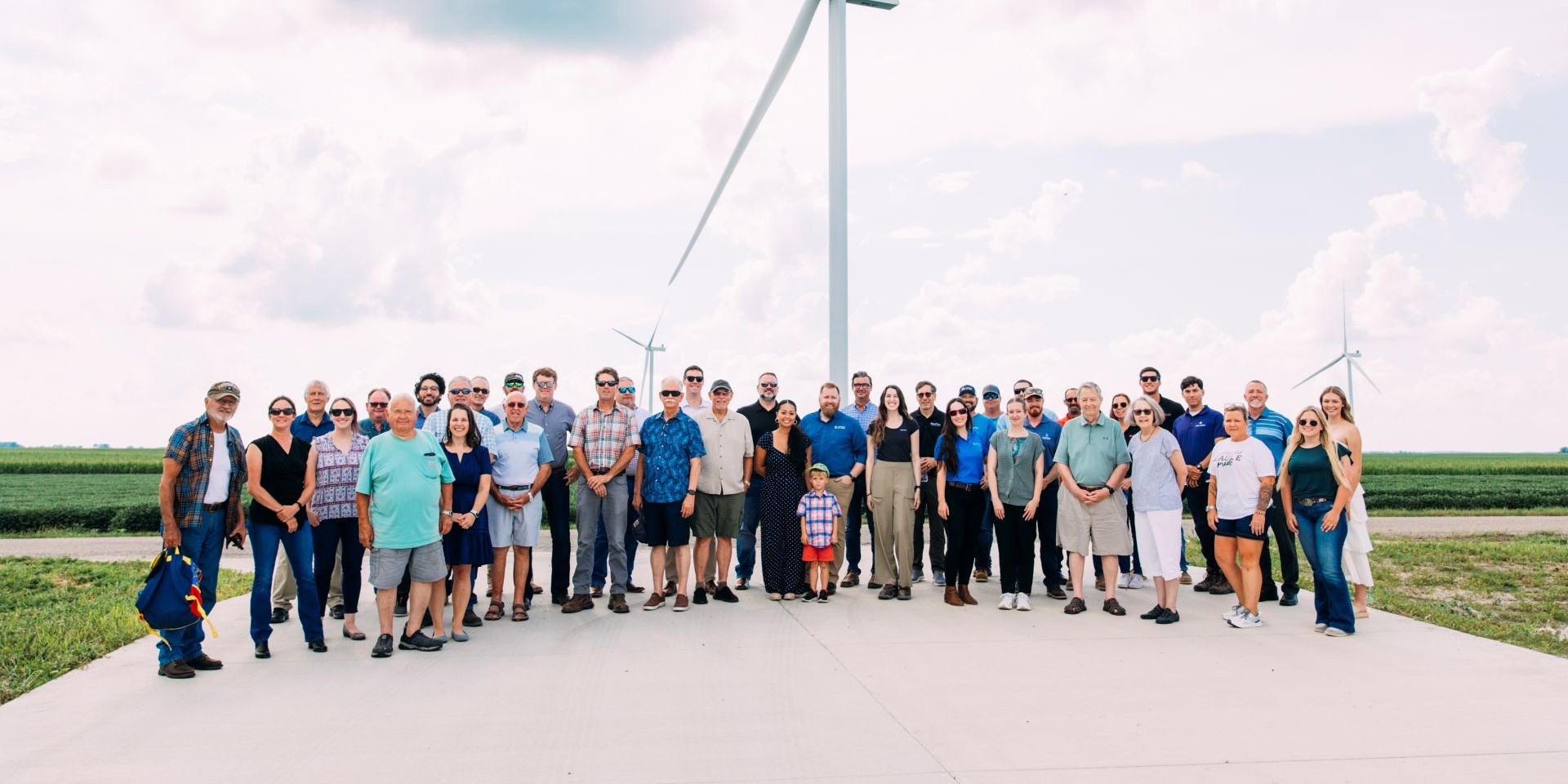 Large group of people of various ages standing outdoors on a concrete surface with wind turbines and green fields in the background under a partly cloudy sky.
