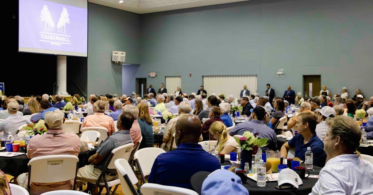 A large audience seated at round tables in a hall attending a Timbermill Wind event, with a screen displaying the Timbermill Wind logo at the front.