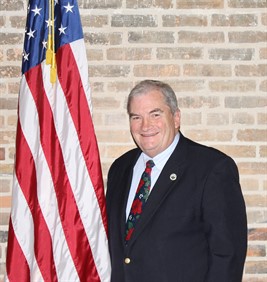 Smiling man in a dark suit jacket, white shirt, and festive tie standing in front of an American flag and a brick wall.