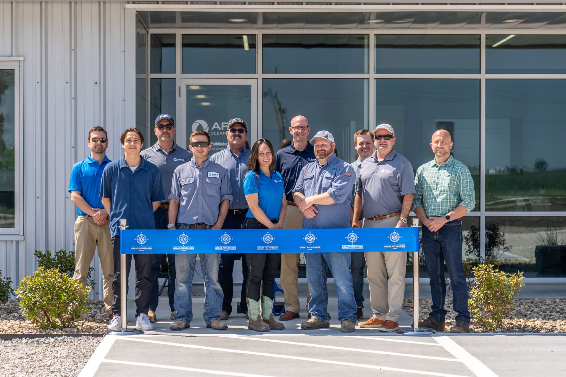 Group of eleven people standing in front of a glass-door building during a ribbon-cutting ceremony. They hold a blue ribbon with the logo and text 'Great Pathfinder' and are dressed in business casual and work attire, with some wearing sunglasses and caps.