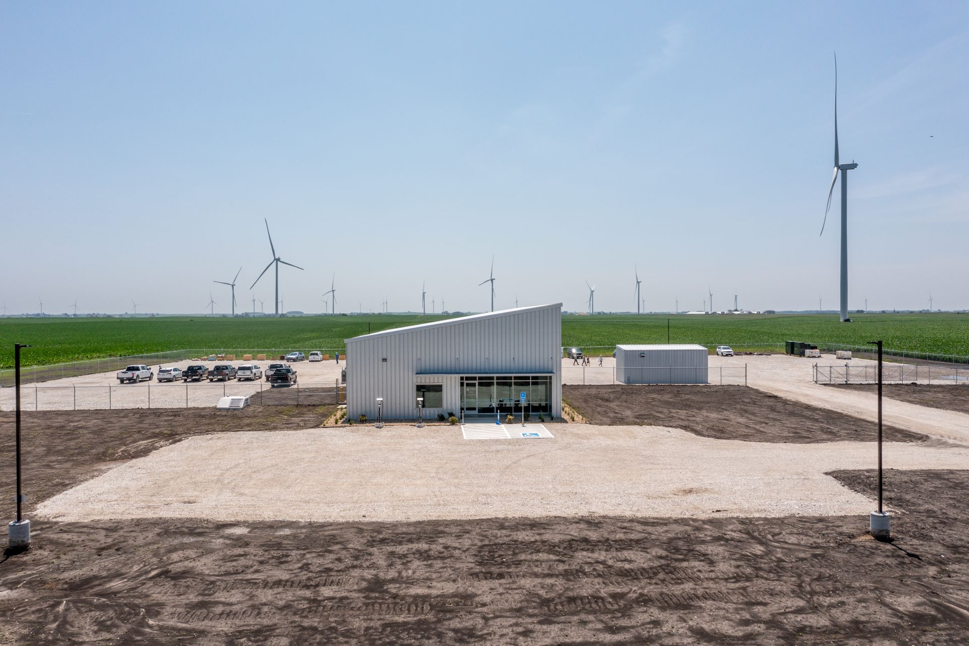 A modern industrial building with slanted roof in the center, surrounded by a fenced gravel parking lot with several cars and a smaller adjacent structure, set against a vast green field with numerous wind turbines under a clear blue sky.