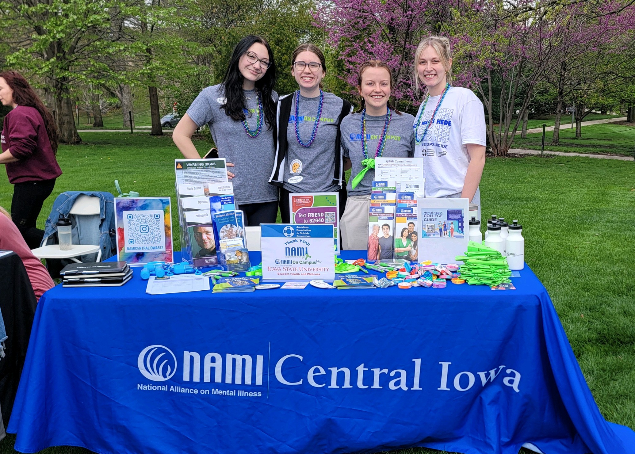 Four smiling women standing behind a table covered with a blue cloth labeled NAMI Central Iowa, National Alliance on Mental Illness, with brochures, buttons, water bottles, and other informational items on mental health, set outdoors with green grass and blooming trees in the background.