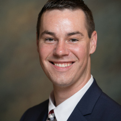 Portrait of a smiling man with short dark hair wearing a dark suit jacket, white shirt, and striped tie, against a neutral blurred background.