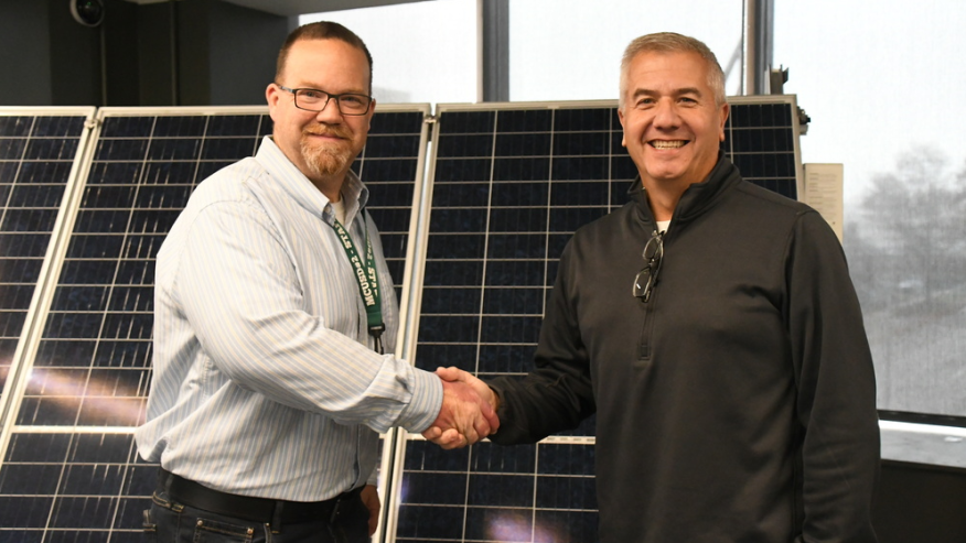 Two men smiling and shaking hands in front of solar panels inside a building with large windows.