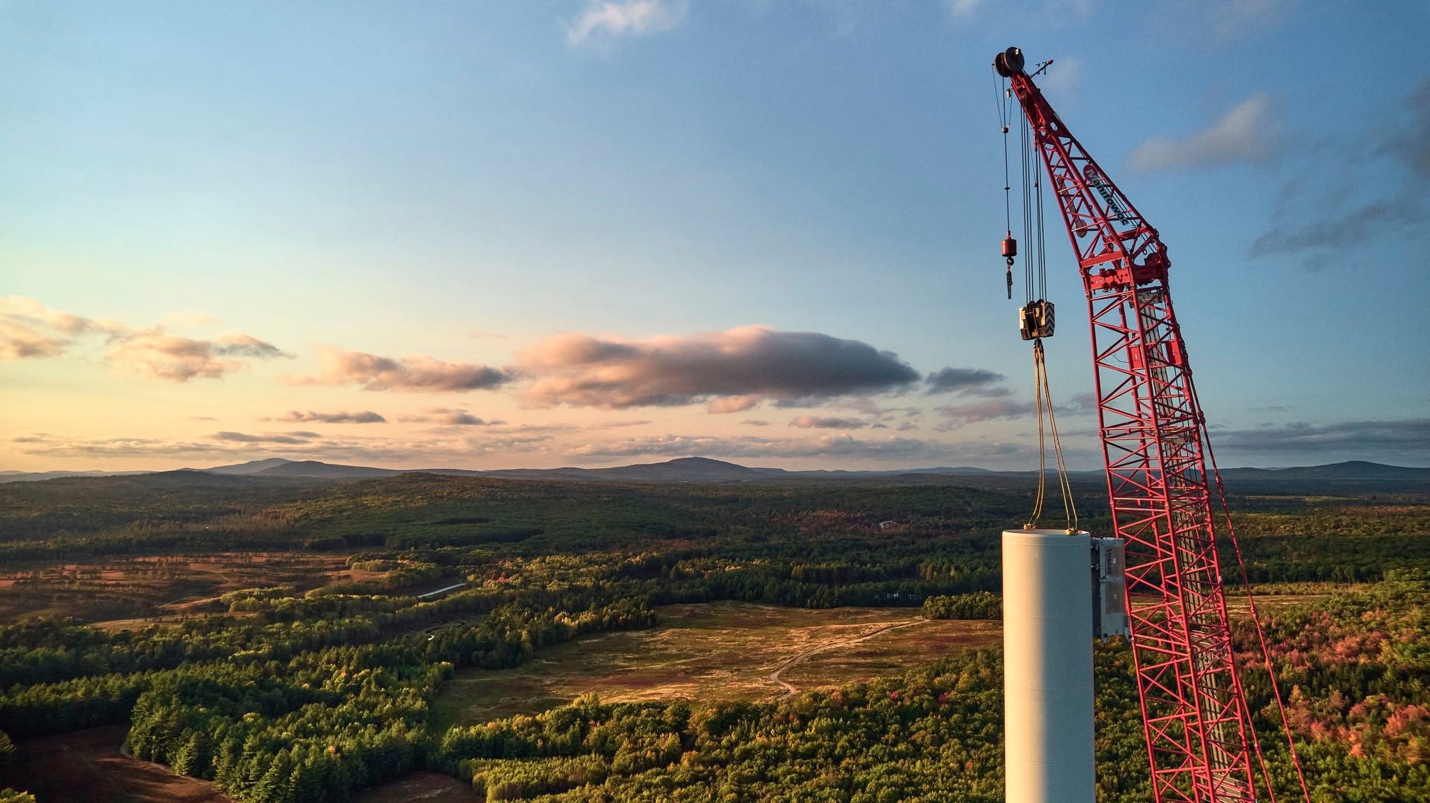 A red crane hoisting the top section of a white wind turbine tower with a forested landscape and hills in the background under a partly cloudy sky at sunset.
