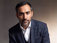 Portrait of a serious man with short dark hair and beard wearing a dark blazer and white shirt.
