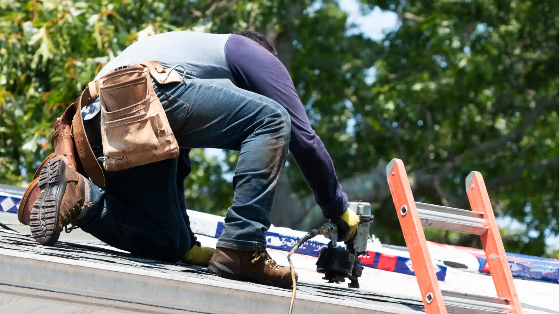 A roofer kneeling on a roof using a nail gun beside an extension ladder