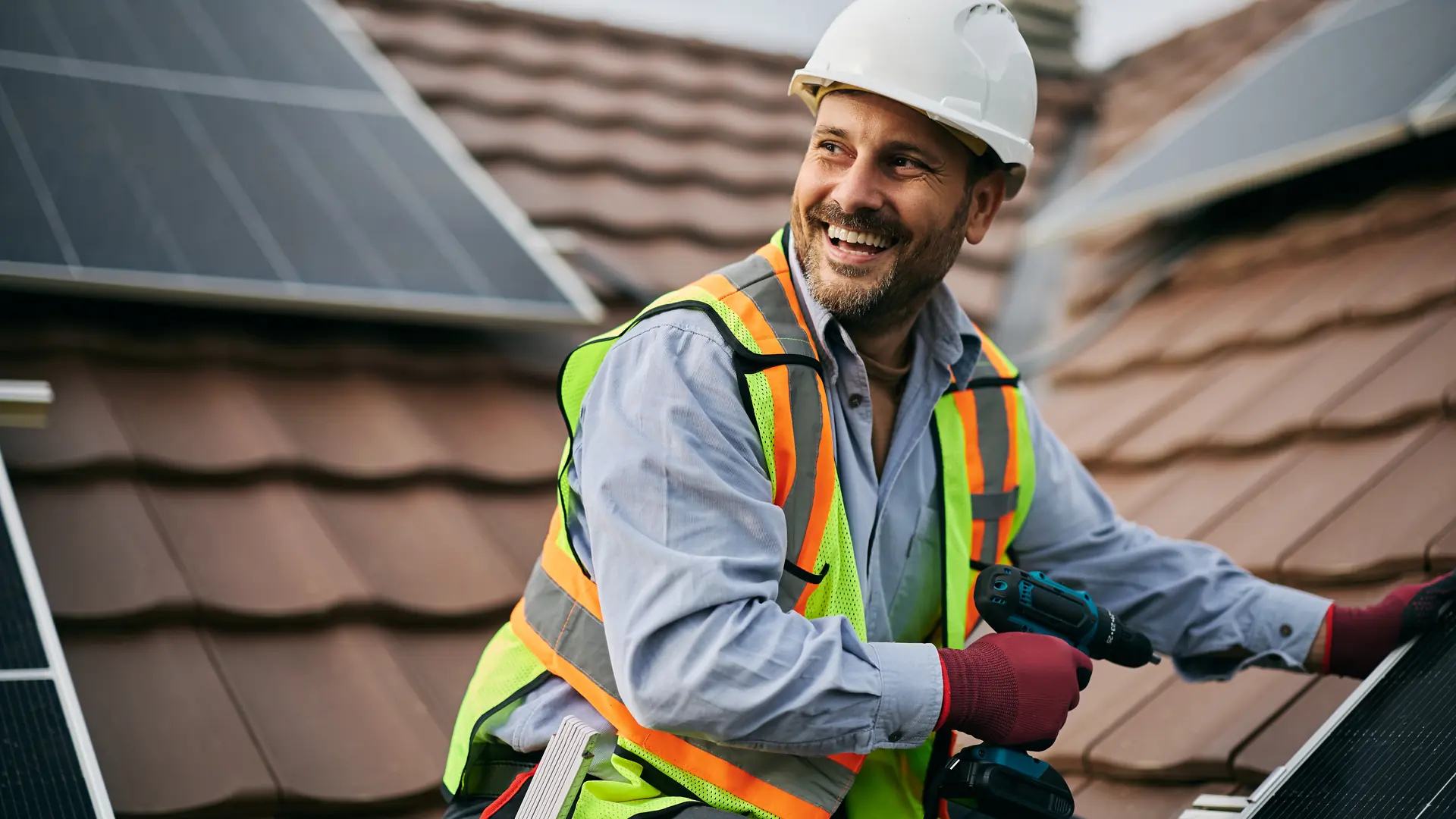 A smiling contractor in a hard hat and high-visibility vest installing solar panels on a tiled roof