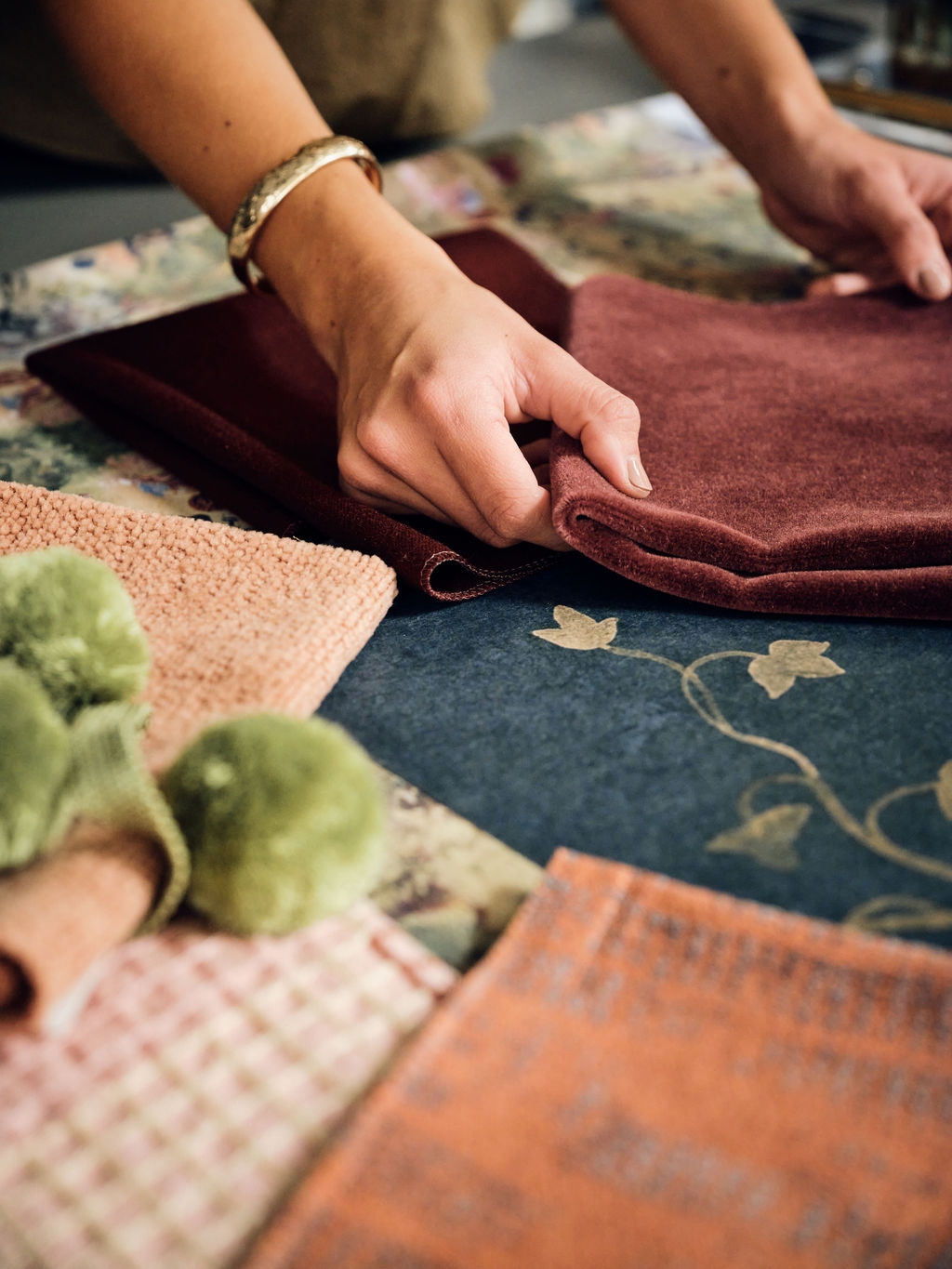 Hands folding a burgundy velvet swatch beside green pom-pom trim