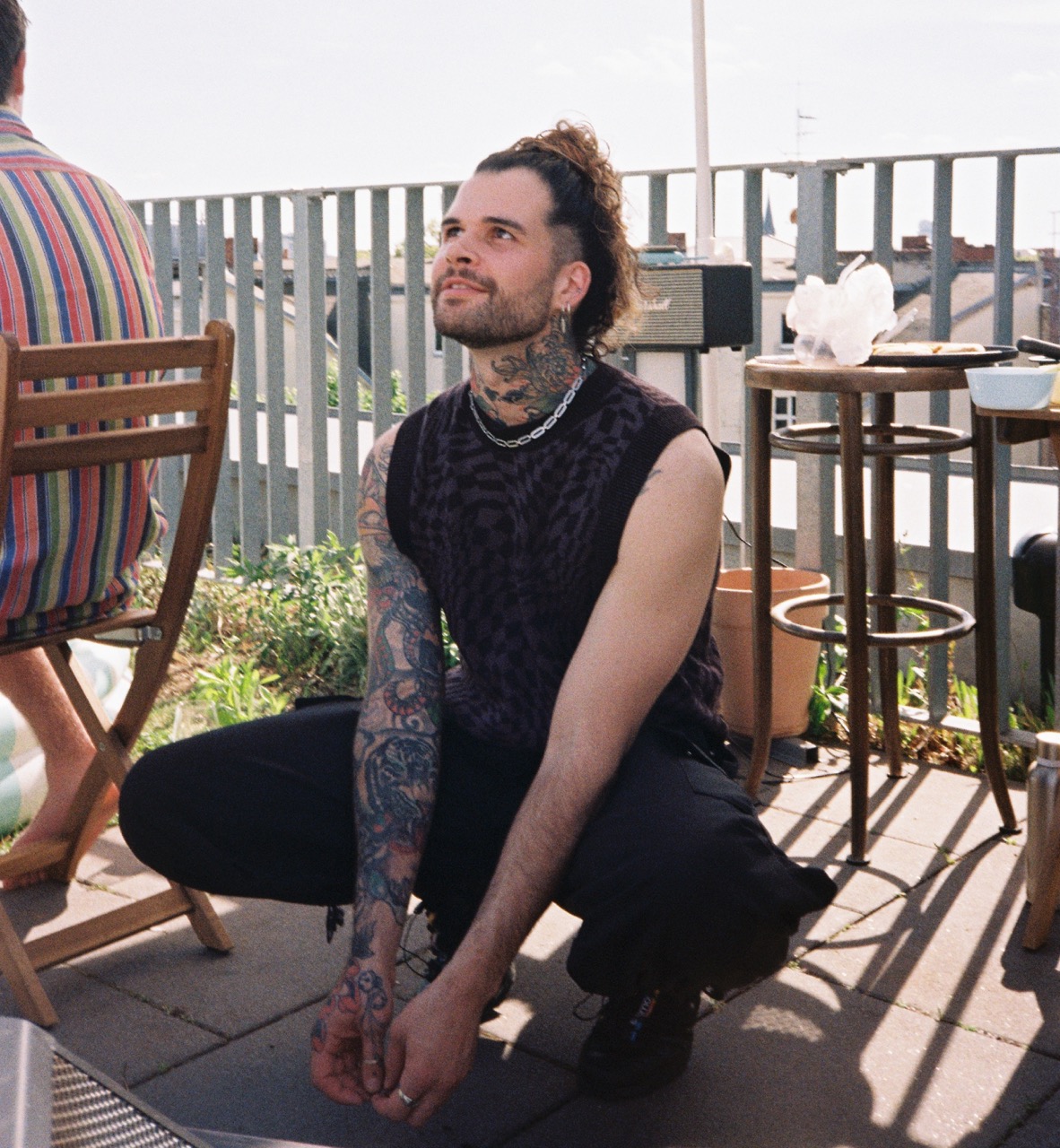 Nikolas Haniotes crouching on a sunlit rooftop terrace, looking up with a slight smile, tattooed arms resting on his knees; a railing, plants and a bar table behind him.