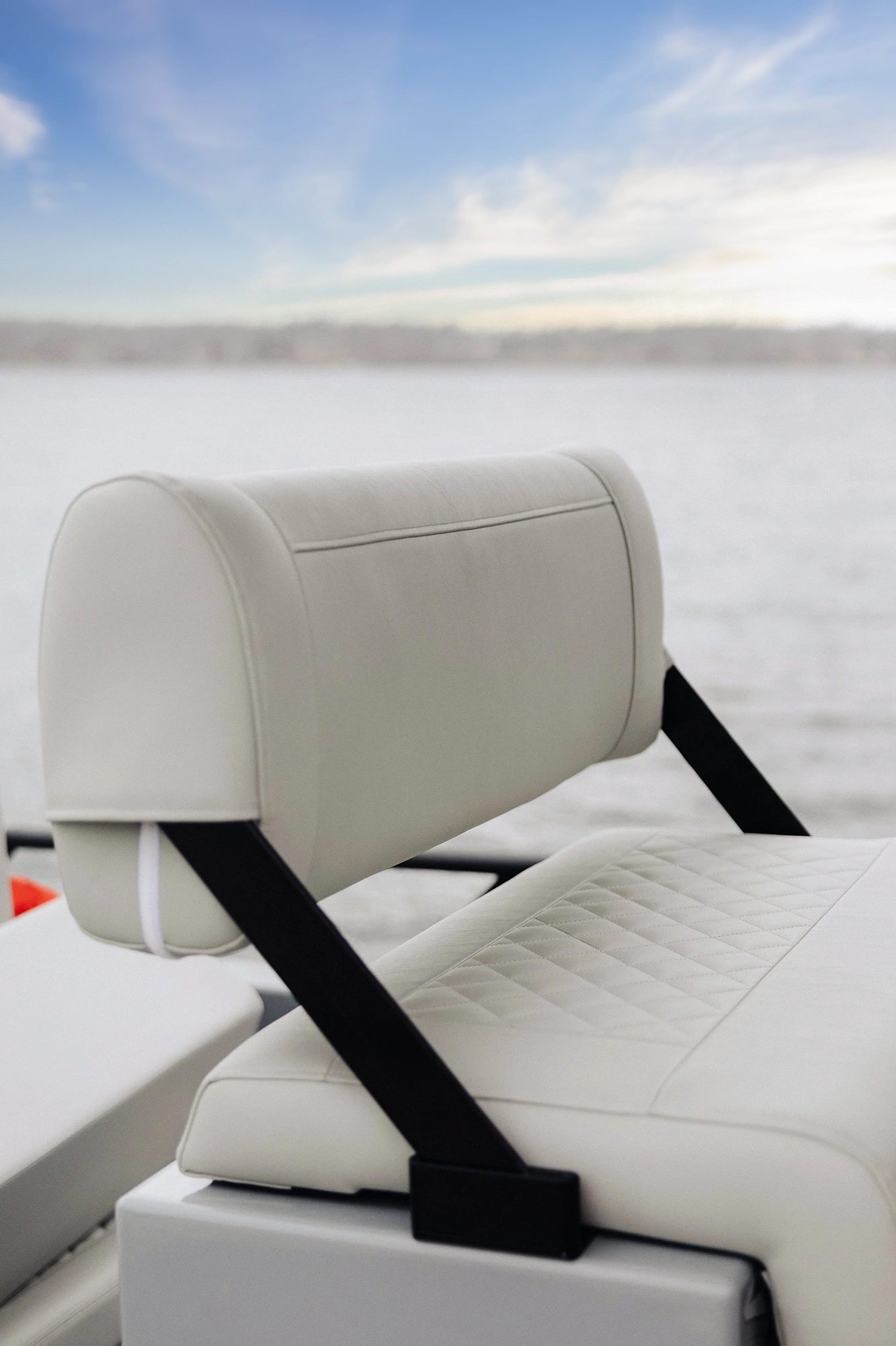 Close-up of a white cushioned boat seat with a quilted pattern on the seat and a curved backrest supported by black straps, with a blurred water background under a partly cloudy sky.