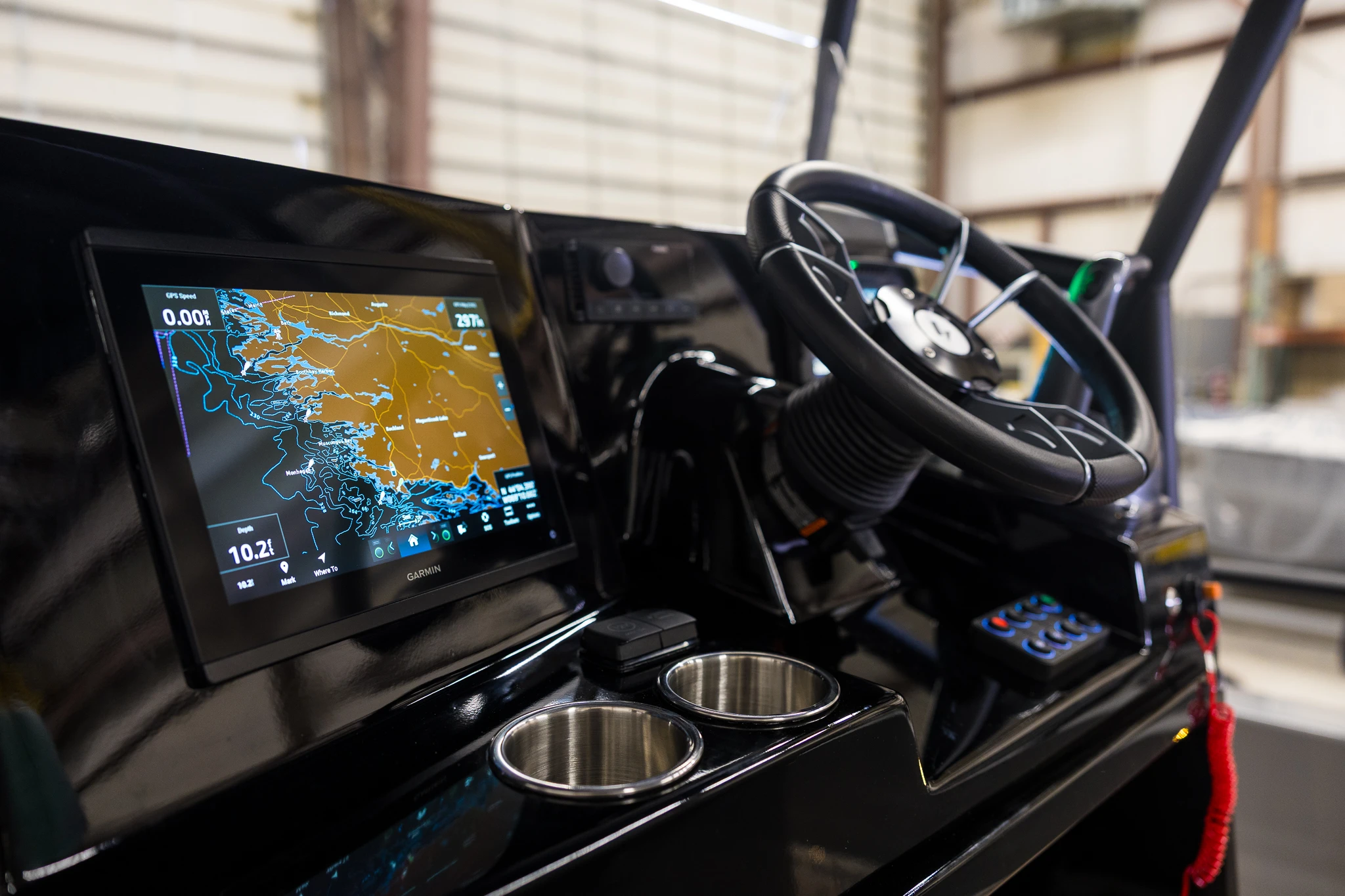 Boat dashboard featuring a Garmin navigation screen displaying a detailed map with GPS speed and depth, a black steering wheel, stainless steel cup holders, control buttons, and part of a red safety lanyard.